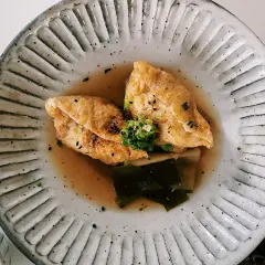 Two pieces of golden aburaage tofu simmer in a savory broth with leeks and seaweed, garnished with green onions and sesame seeds.