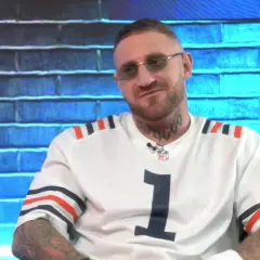 Mężczyzna w okularach i białej koszulce z numerem 1 na tle niebieskiej ściany z cegły.
