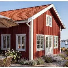 Rött trähus vid havet med vita fönster och en dropplist i fasaden. En segelbåt syns i bakgrunden.