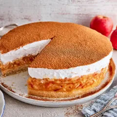 Eine köstliche schwedische Apfeltorte mit einer luftigen Sahnehaube, bestäubt mit Zimt. Daneben liegen frische Äpfel.