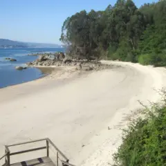 Escaleras de madera bajan a la playa Niño do Corvo, con arena blanca, rocas y mar azul.
