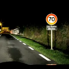 Route de nuit avec marquage au sol blanc lumineux. Panneau "Les Quatre Moulins" et limite de vitesse à 70.