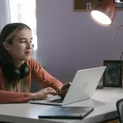 Młoda kobieta z słuchawkami na szyi pracuje na laptopie, być może instaluje aplikację z nieznanych źródeł, ryzykując bezpieczeństwo danych.