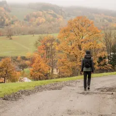 Osoba z plecakiem idzie drogą przez jesienny krajobraz. W oddali widać wzgórza pokryte drzewami w barwach ożennej.