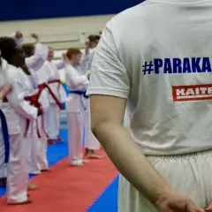 En grupp utövare i vita dräkter tränar karate. En person bär en t-shirt med texten #PARAKARATE och KAITEN.
