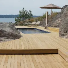 En stor altan i trä med pool och solstolar, omgiven av klippor och med utsikt över vattnet. Trall mått är perfekt för detta sommarparadis.