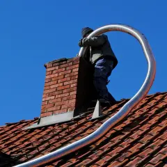 Un couvreur effectue l'installation tubage cheminée sur un toit en tuiles rouges, sous un ciel bleu éclatant.