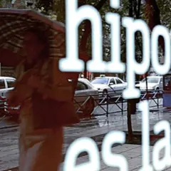 Reflejo en un escaparate: "Hipoteca, ¿es la fein suya?". Una persona con paraguas pasa por la calle lluviosa con taxis.