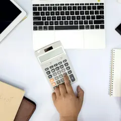 Ręka naciska przycisk kalkulatora, by sprawdzić, co to jest saldo rachunku. Obok leży laptop, tablet i notatnik.