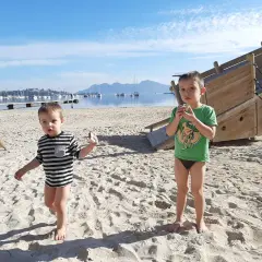 Dwóch chłopców na plaży, cieszących się słoneczną, majową pogodą w lutym. W tle widać morze z łodziami i góry.