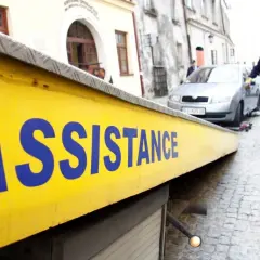 Żółta platforma lawety z napisem "ASSISTANCE" w niebieskich literach. W tle mężczyzna w jaskrawej kamizelce pomaga przy samochodzie.