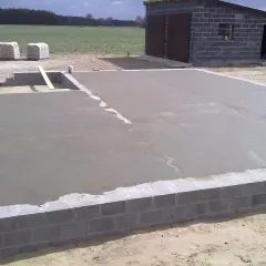 Świeżo wylana płyta betonowa, z widoczną minimalną grubością chudziaka, na fundamencie z bloczków. W tle budynek gospodarczy.