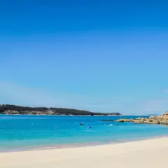 Arena blanca y aguas turquesas en la playa de Menduiña, con barcas a lo lejos y una costa rocosa.