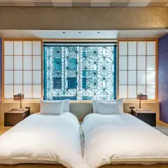 Dwie białe pościele w pokoju hotelowym w stylu japońskim. W tle okno z geometrycznym wzorem i shoji.