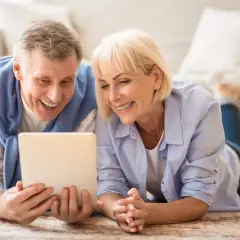 Ein lachendes älteres Paar liegt auf dem Boden und schaut auf ein Tablet. Ihre positiven senioren dates erfahrungen werden durch ihr Lächeln deutlich.