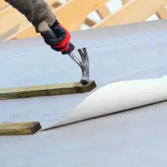 Ręka w rękawicy przybija młotkiem drewnianą łatę do dachu pokrytego membraną.