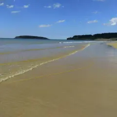 Arena dorada y mar tranquilo en la playa de Loredo, Cantabria. El sol brilla en un cielo azul con nubes dispersas.