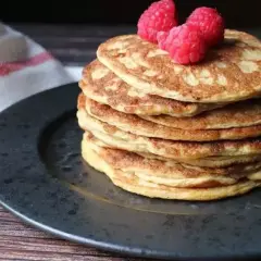 En hög med nyttiga pannkakor toppas med hallon, serverade på en mörk tallrik med en vit handduk med röda ränder i bakgrunden.