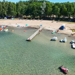 Słoneczny dzień nad jeziorem, gdzie kolorowe rowerki wodne i kajaki czekają na plaży. W tle widać namioty i kampery, tworząc atmosferę letniego binduga camping.