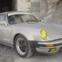 Srebrny Porsche 911 z żółtymi reflektorami, w stylu cyberpunk, zaparkowany na tle zniszczonej ściany z graffiti.