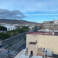 Vista aérea de una ciudad con edificios, palmeras y montañas al fondo. Las hortichuelas se ven en la distancia, bajo un cielo nublado.