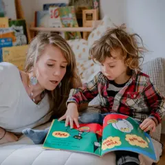 Kobieta czyta dziecku książkę, pokazując ilustracje. W tle regały z książkami, co oddaje atmosferę przytulnego miejsca do nauki i zabawy, jak wygląda żłobek.