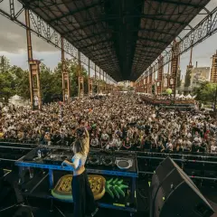 DJ sul palco, folla immensa sotto una struttura industriale, un'atmosfera da festival con il logo "Kappa" visibile.