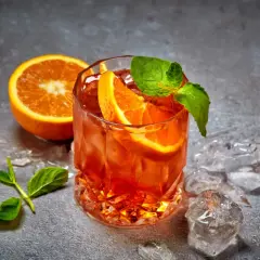 Un cocktail aranciato con fette d'arancia e menta, circondato da cubetti di ghiaccio per cocktail che non si scioglie, su uno sfondo grigio.