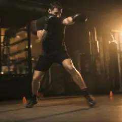 A determined pivot fighter in black boxing gloves and shorts practices in a dimly lit gym, his body a blur of motion.