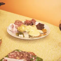 Un verre de vin rouge accompagne une raclette savoureuse, des charcuteries et des fromages sur une table jaune.
