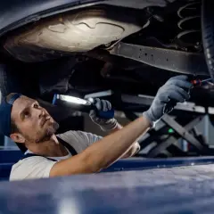 Mechanik z latarką bada podwozie auta. Sprawdza, czy potrzebna jest wymiana wahacza, cena usługi zależy od modelu.