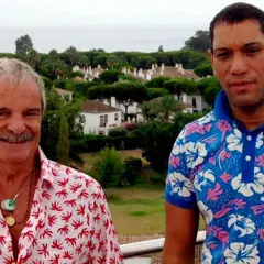 Elio Valderrama Prescott y un amigo posan con camisas coloridas frente a un paisaje de casas y árboles.