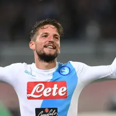 Uśmiechnięty Dries Mertens w białej koszulce Napoli z rękami uniesionymi w geście radości.