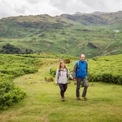 A couple enjoys hiking in England, hand-in-hand, amidst lush green hills and ferns under a cloudy sky.