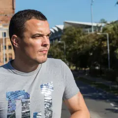 Dawid Janczyk w szarej koszulce z błyszczącym napisem, na tle miejskiego krajobrazu z budynkami i drzewami.