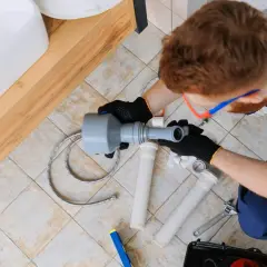Instalator naprawia rury pod umywalką, prawdopodobnie z powodu braku odpowietrzenia pionu.