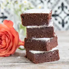 Schokoladige Brownies mit Datteln, bestäubt mit Puderzucker, neben einer zarten orangefarbenen Rose auf Holztisch.