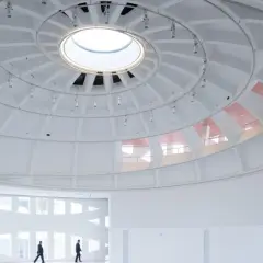 The expansive, white interior of the Faena Art Project Room features a striking domed ceiling with a central skylight. Two figures walk through the minimalist space.
