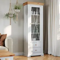 Jasny salon biało drewniany z witryną pełną szkła i porcelany. Wiszące makramy z roślinami dodają przytulności.