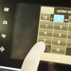 Palec dotyka ekranu podczas ustawiania godziny w Opel Astra H. Wyświetlacz pokazuje klawiaturę numeryczną do wprowadzania czasu.