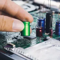 Dłoń trzyma zielony kondensator, przygotowując go do montażu na płycie głównej. W tle widać inne kondensatory i układy elektroniczne.