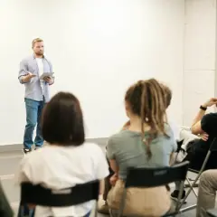 Mężczyzna z tabletem przemawia na zebraniu wspólnoty mieszkaniowej. Słuchacze siedzą na krzesłach, skupieni na prelegencie.
