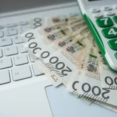 Kalkulator i banknoty 200 zł na klawiaturze laptopa. Obrazek sugeruje analizę finansową, być może związaną z grupami podatkowymi.