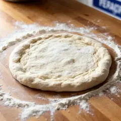 Pâte à pizza qui colle, étalée sur un plan de travail en bois, saupoudrée de farine.