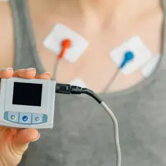 Dłoń trzyma urządzenie do monitorowania ekg serca. Na klatce piersiowej widoczne elektrody.