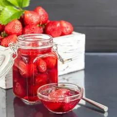 Słoik i miseczka z kompotem z truskawek, obok skrzynka pełna świeżych owoców. Idealny przepis na to, jak zrobić kompot z truskawek.