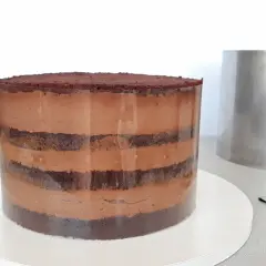 Czekoladowa torta z warstwami ciasta i kremu, przygotowywana w transparentnej formie. Obok stoi metalowy pierścień.