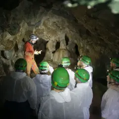 Exploradores en la cueva Rei Cintolo, con cascos verdes y trajes blancos, escuchan atentamente a su guía.
