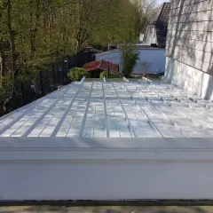 Neues, weißes Metalldach mit sichtbarem Ortgang. Im Hintergrund ein kleiner Garten mit Bäumen und einem roten Gartenhaus.