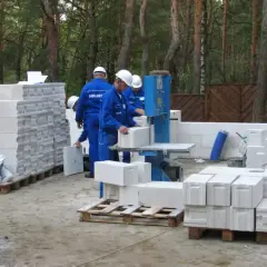 Budowa domu z bloczków betonowych. Pracownicy w niebieskich kombinezonach i białych kaskach przygotowują bloczki, sprawdzając ich bloczek betonowy wymiary.